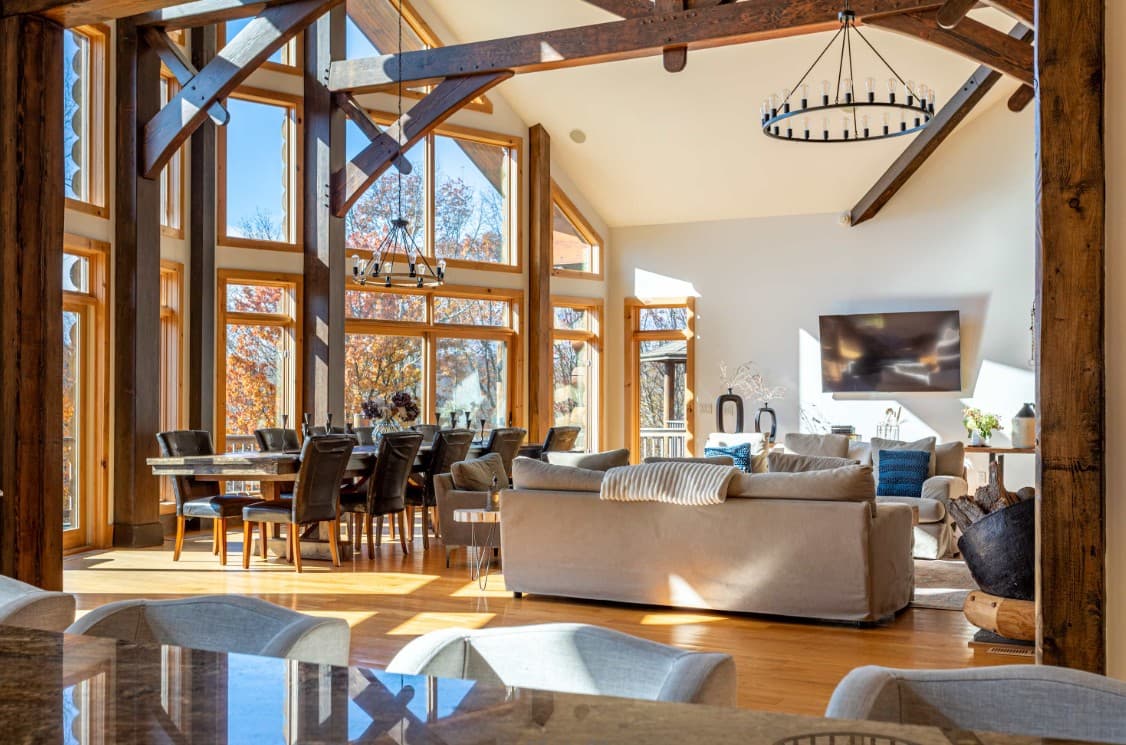 Bright great room with dining table, sofas, and floor-to-ceiling forest views